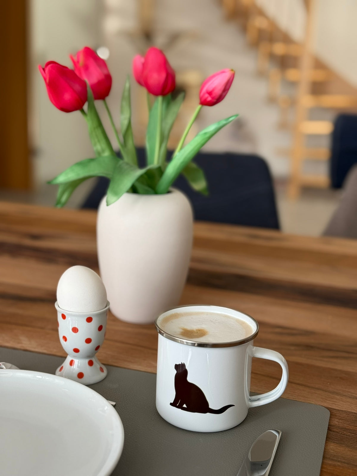 Emaille-Tasse mit Kaffee auf einem modern gedeckten Esstisch mit Tulpen und Eierbecher.
